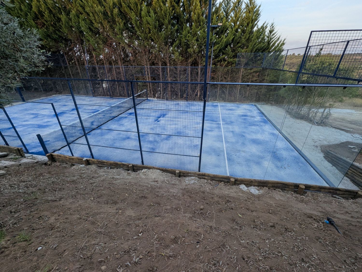 O novo campo de padel: piso azul, estrutura azul-escura e vidros, rodeado de ciprestes
